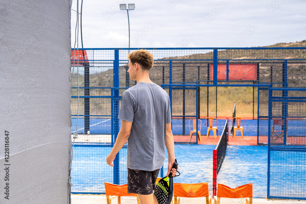 Rear view of a young player carrying a padel racket and stepping into the court, ready for an exciting game under a clear sky.