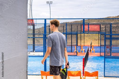 Rear view of a young player carrying a padel racket and stepping into the court, ready for an exciting game under a clear sky.