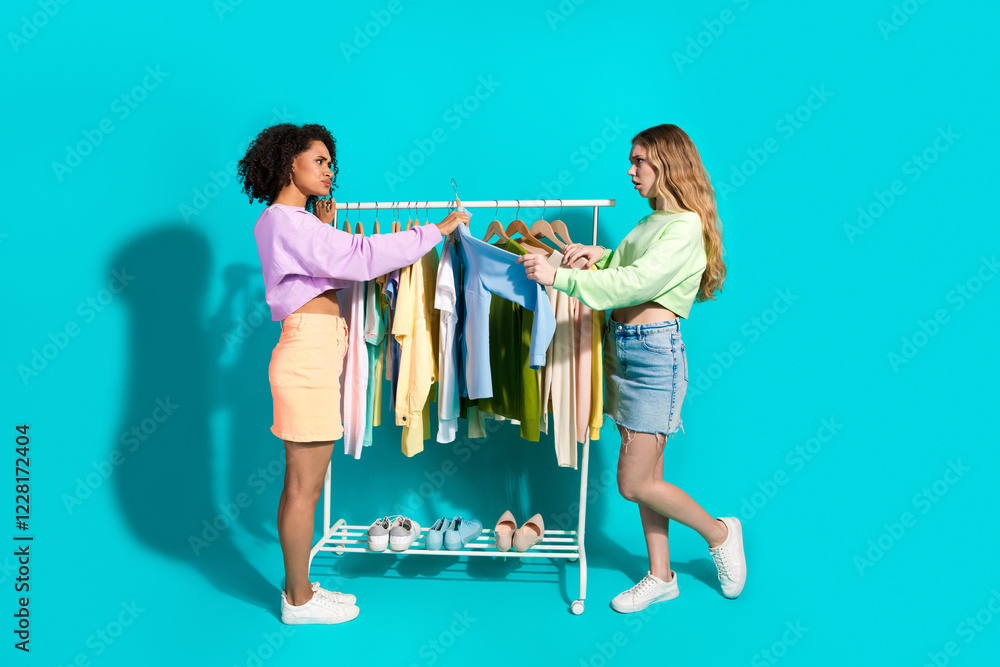 Naklejka premium Two young women interact over a clothing rack in a vibrant turquoise setting, showcasing fashion, youth, and a stylish casual relationship.