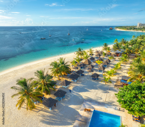 Fototapeta Naklejka Na Ścianę i Meble -  Aerial view of pool, white sandy beach with palms, umbrellas, boat, blue sea at sunset. Summer vacation in Nungwi, Zanzibar island. Tropical landscape. Clear azure sea. Top drone view of luxury resort