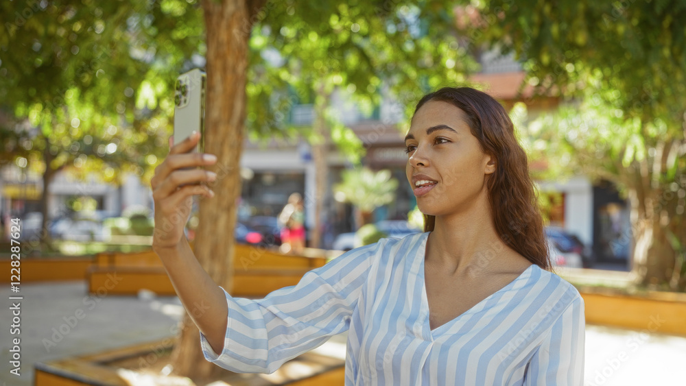 Naklejka premium Woman taking selfie outdoors in city park during daytime, brunette and beautiful in contemporary urban setting with trees and buildings.
