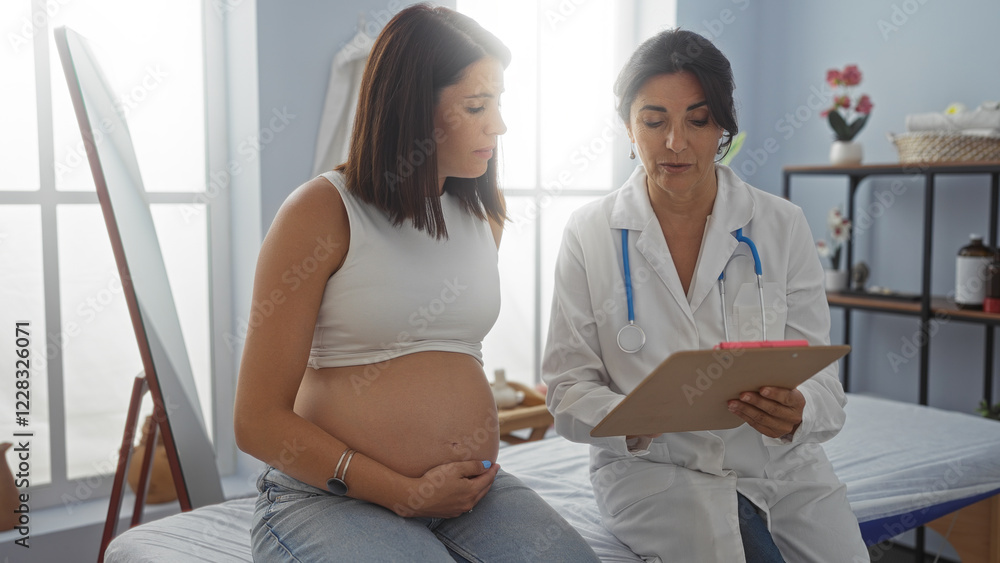 Obraz premium Pregnant woman sits with female doctor in a medical clinic room, discussing pregnancy details and health while holding clipboard.