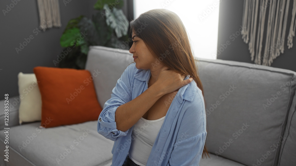 Young woman experiencing neck pain in a living room setting, with her hand on her neck and a pained expression, seated on a gray sofa near cushions and plants Young woman experiencing neck pain in a living room setting, with her hand on her neck and a pained expression, seated on a gray sofa near cushions and plants