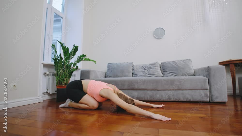 A woman is performing the downward dog yoga pose in a spacious living room with natural light. The room features a sofa and indoor plants. A woman is performing the downward dog yoga pose in a spacious living room with natural light. The room features a sofa and indoor plants.