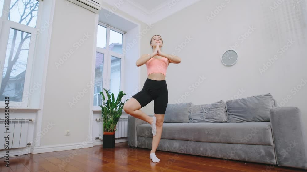 A woman balances on one leg while practicing yoga in a well-lit living room with a cozy atmosphere. Green plants add freshness to the space. A woman balances on one leg while practicing yoga in a well-lit living room with a cozy atmosphere. Green plants add freshness to the space.