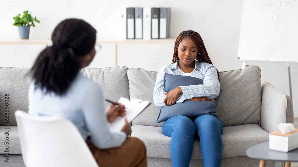 Depressed black woman sitting on couch, hugging pillow, having consultation with psychologist at modern clinic. Treatment of anxiety, emotional and mental disorders concept Depressed black woman sitting on couch, hugging pillow, having consultation with psychologist at modern clinic. Treatment of anxiety, emotional and mental disorders concept