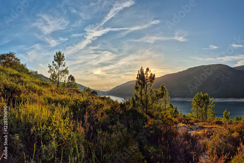 Golden sunlight casts a warm glow over Vilarinho das Furnas Reservoir in Peneda-Gerês National Park, Portugal. A serene landscape with rolling hills, lush vegetation, and calm waters reflecting the sk photo