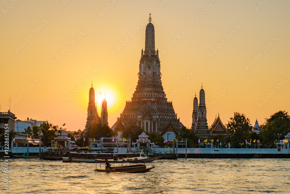 Fototapeta premium A stunning sunset envelops Wat Arun, its intricate spires silhouetted against the golden sky. Local boats softly glide along the Chao Phraya River, creating a serene atmosphere in vibrant Bangkok.