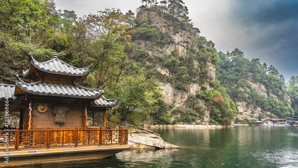 Fototapeta premium The beautiful lake is surrounded by steep picturesque mountains. Green vegetation on the slopes of the cliffs. In the foreground is a Chinese boat with curved roofs and carved wooden railings. China.
