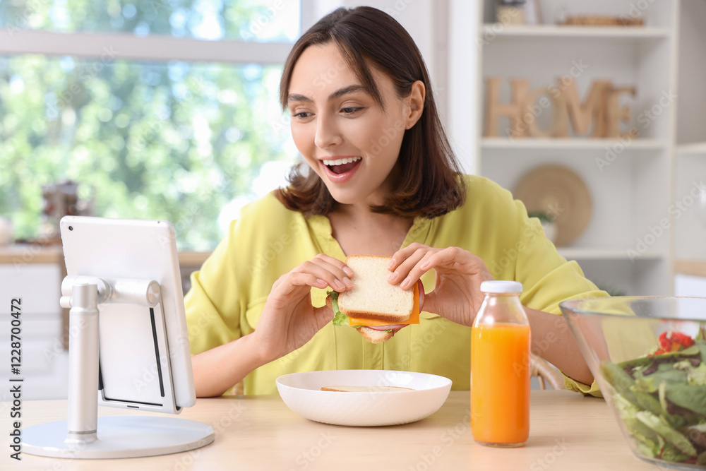 Beautiful young woman eating tasty sandwich while watching something on tablet computer in kitchen Beautiful young woman eating tasty sandwich while watching something on tablet computer in kitchen