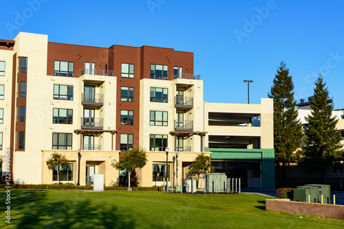 Modern multi-story apartment building with beige and brown exterior, balconies, adjacent to a parking garage, and a manicured lawn in the foreground