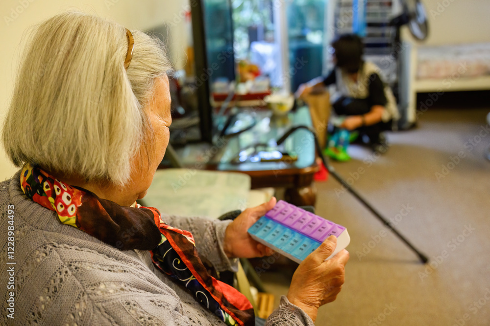 Senior woman holding a pill box inside home. Out-of-focus adult daughter cleaning the house.