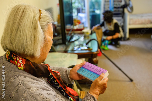 Obraz na płótnie Senior woman holding a pill box inside home. Out-of-focus adult daughter cleaning the house. w salonie