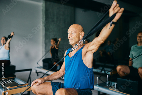 Fit senior man in sportswear focused on doing resistance training on a reformer during a class with other people in a pilates gym photo