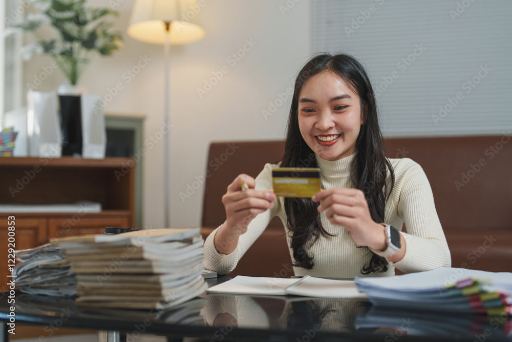 Young woman sits at a table, smiling as she holds a credit card. Stacks of paperwork and a cozy interior setting suggest financial planning or online shopping activities Young woman sits at a table, smiling as she holds a credit card. Stacks of paperwork and a cozy interior setting suggest financial planning or online shopping activities
