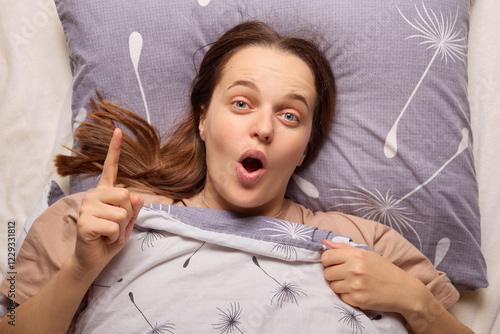 A woman lying in bed looks surprised with her mouth open and a hand raised as if sharing an idea. She is under a soft blanket, surrounded by decorative pillows photo