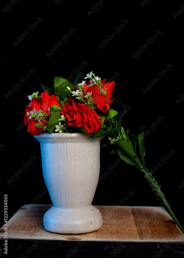 Fresh red roses with green leaves and white blossoms beside an empty white vase, set on a wooden surface against a black background. Fresh red roses with green leaves and white blossoms beside an empty white vase, set on a wooden surface against a black background.