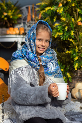 A beautiful Russian girl in a fur coat and headscarf sits on the porch of a wooden house on a sunny spring day. Family values, traditions and home comfort.Tea drinking outside the house