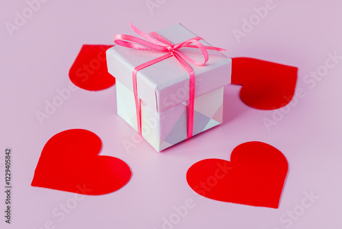 A gift box tied with a red ribbon with a bow on a pink background with red hearts. Valentine's Day gift concept.