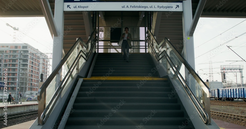Obraz premium A woman in formal clothes and headphones, with a cup of coffee in her hand, descends the stairs to the platform of the railway station