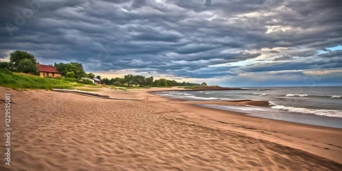 Obraz Sandy beach meets rolling waves under a dramatic cloudy sky with a cozy house nestled nearby