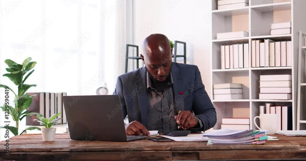 Man of successful African American businessman in elegant suit using laptop gadgets while sitting by workplace in front of documents. Man of successful African American businessman in elegant suit using laptop gadgets while sitting by workplace in front of documents.