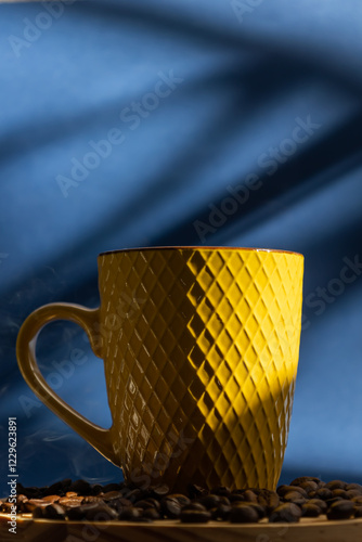 Vertical image of steaming coffee in a yellow cup on a wooden plate with scattered coffee beans on a blue background with sun rays coming through the window photo