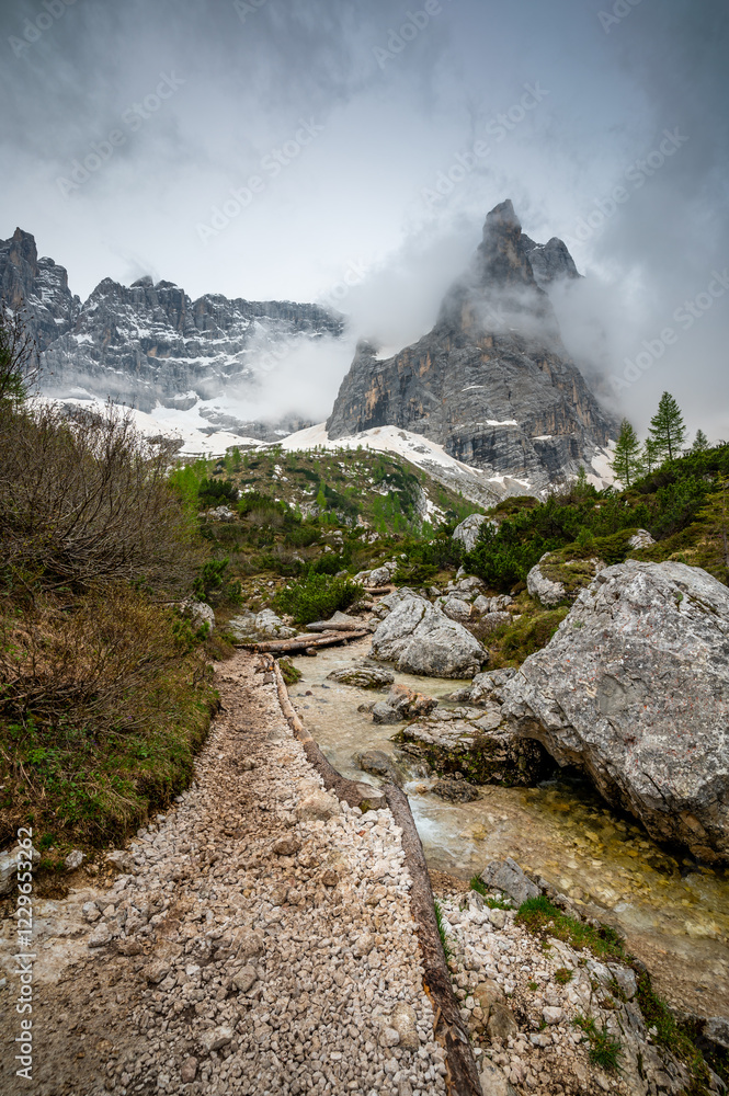 Rocky hiking trail near Lago Sorapis in the Dolomites (Dolomiti, Dolomiten), Italy. The path runs alongside a small mountain stream, with massive boulders and peaks in the background