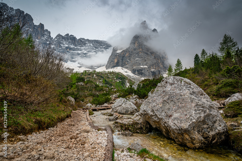 Rocky hiking trail near Lago Sorapis in the Dolomites (Dolomiti, Dolomiten), Italy. The path runs alongside a small mountain stream, with massive boulders and peaks in the background