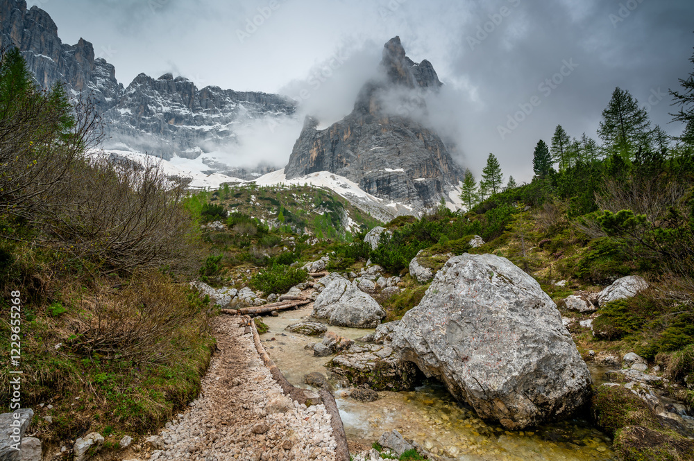 Rocky hiking trail near Lago Sorapis in the Dolomites (Dolomiti, Dolomiten), Italy. The path runs alongside a small mountain stream, with massive boulders and peaks in the background