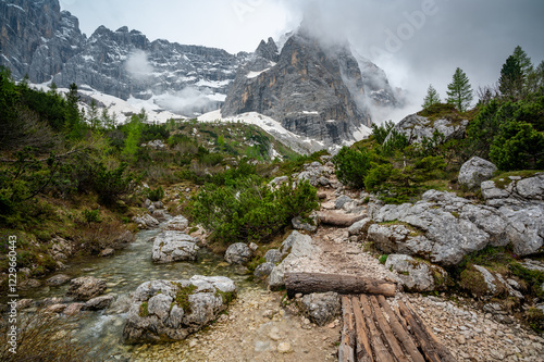 Rocky hiking trail near Lago Sorapis in the Dolomites (Dolomiti, Dolomiten), Italy. The path runs alongside a small mountain stream, with massive boulders and peaks in the background