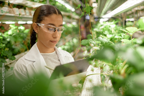 Young woman wearing protective eyewear studying plants in indoor cultivation environment with multi-level shelving under artificial lighting photo