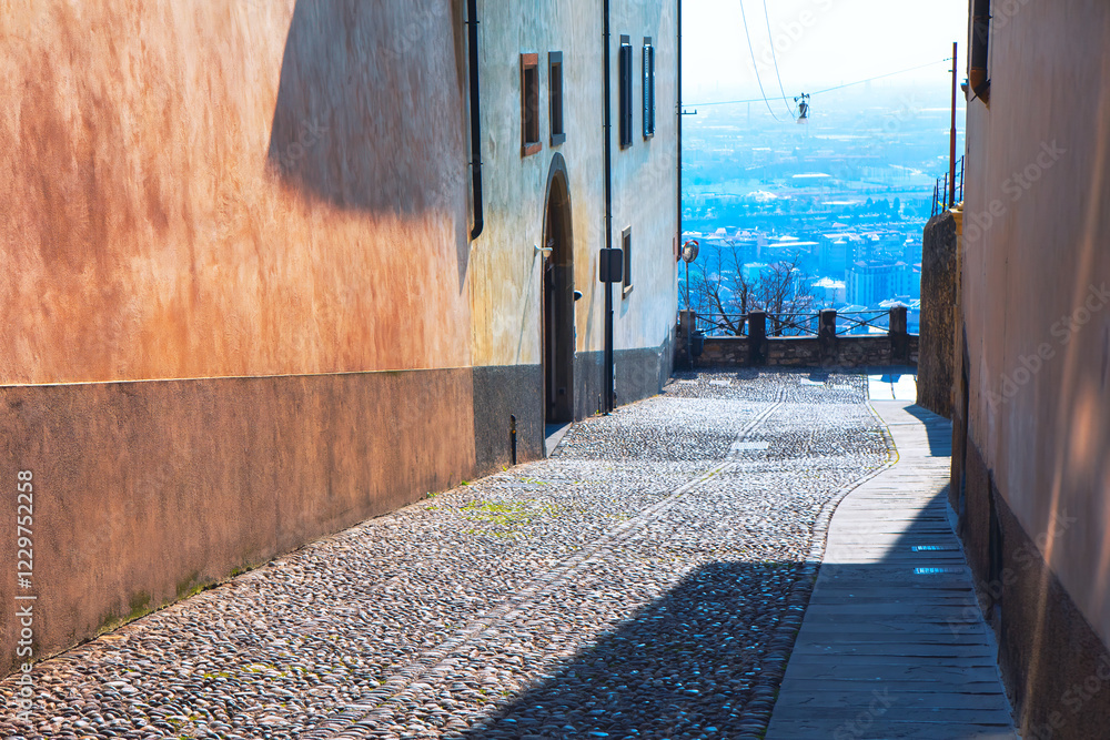 Narrow cobblestone alleyway flanked by tall buildings with a view of a distant cityscape in background. Alleyway is partially shaded, with a stone pathway
