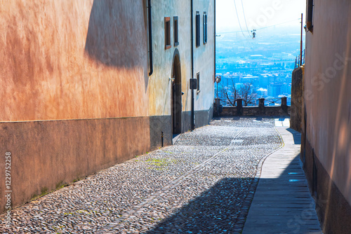 Obraz na płótnie Narrow cobblestone alleyway flanked by tall buildings with a view of a distant cityscape in background. Alleyway is partially shaded, with a stone pathway w salonie