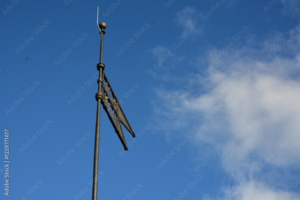 A metal weather vane with an intricate design stands atop a pole against a clear blue sky. The decorative element appears to have a pattern resembling a bird or emblem A metal weather vane with an intricate design stands atop a pole against a clear blue sky. The decorative element appears to have a pattern resembling a bird or emblem