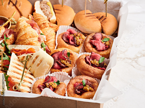 A variety of delectable savory treats is displayed in a well-organized box, featuring sandwiches, pastries, and meats garnished with herbs, perfect for any occasion photo