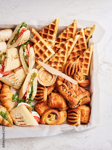 A vibrant spread showcases assorted sandwiches with fresh vegetables, golden waffles, and a selection of flaky pastries arranged beautifully. Perfect for a brunch gathering photo