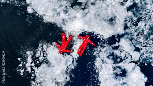 Aerial view of two people ice floating on icy water in bright red survival suits. Arctic frozen landscape. Extreme adventure, ice diving, cold water survival, winter expedition. Fun, outdoor activity