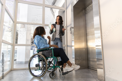 Disabled woman in wheelchair presses waiting the elevator with female colleague in the office