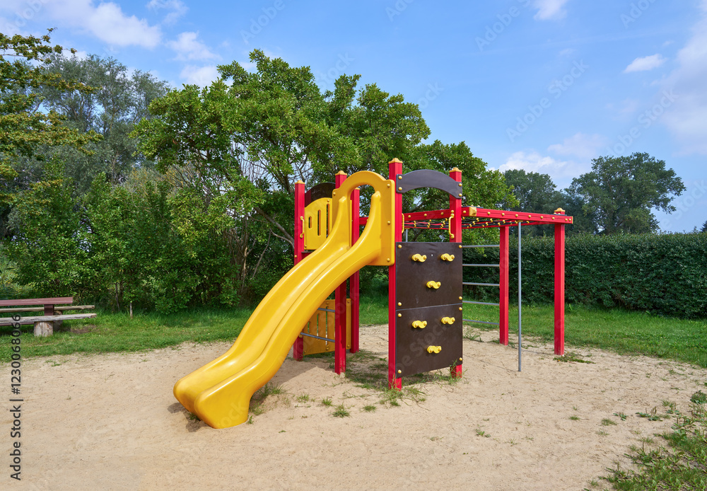 Farbiges Klettergerüst mit Rutsche auf einer Sandfläche - Kinderspielplatz umgeben von Hecken und Bäumen im Sommer Farbiges Klettergerüst mit Rutsche auf einer Sandfläche - Kinderspielplatz umgeben von Hecken und Bäumen im Sommer