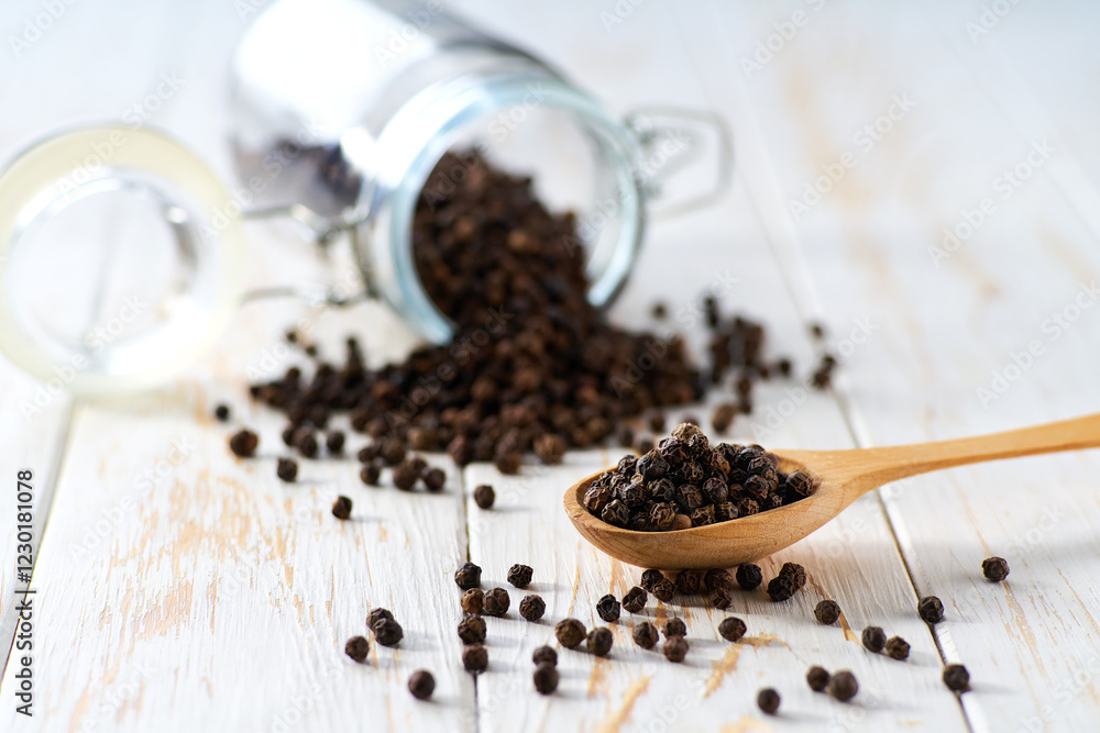 black peppercorns in a wooden scoop and in glass storage jar on a light kitchen table, selective focus. black peppercorns in a wooden scoop and in glass storage jar on a light kitchen table, selective focus.