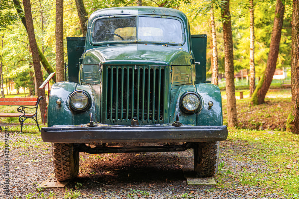 Old green vintage truck parked outdoors in a wooded area, surrounded by trees and a rustic bench.