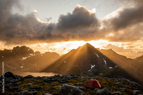 ein kleines oranges zelt steht vor unglaublicher berglandschaft, die sonnenstrahlen kommen durch die wolken