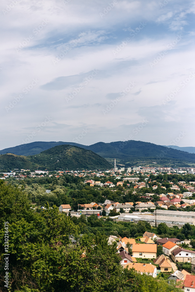 Obraz premium Scenic vista showcasing a cityscape nestled amidst lush greenery, with rolling hills stretching into the distance under a cloudy sky, offering a tranquil and picturesque view