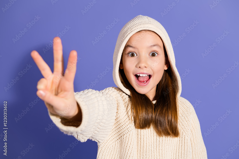 Charming young girl in a casual hoodie smiling joyfully against a purple background, holding up peace sign Charming young girl in a casual hoodie smiling joyfully against a purple background, holding up peace sign