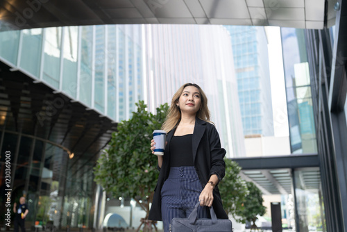 Obraz na plátně A confident young professional woman walking outdoors in a modern urban setting, holding a coffee cup, surrounded by greenery and glass buildings, exuding elegance and contemporary style
