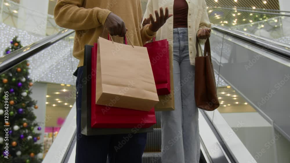 Tilt low angle shot of happy African American couple in casual clothes holding lots of paper bags and talking while going down escalator in shopping mall, discussing purchases, leisure time