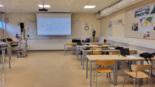 The classroom features multiple desks arranged for students with a projector screen at the front. The space is well-lit and prepared for an upcoming lesson with educational materials around.