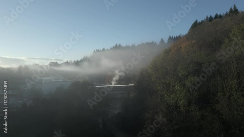 White smoke rises from a factory chimney near a forested hillside, creating a misty layer over sant hilari sacalm in catalonia on a clear morning photo