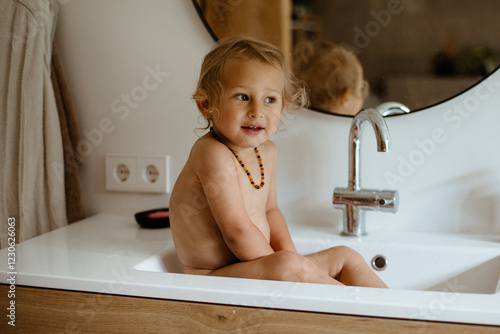 A curly-haired, blond little boy is sitting in the sink, washing himself, and smiling.